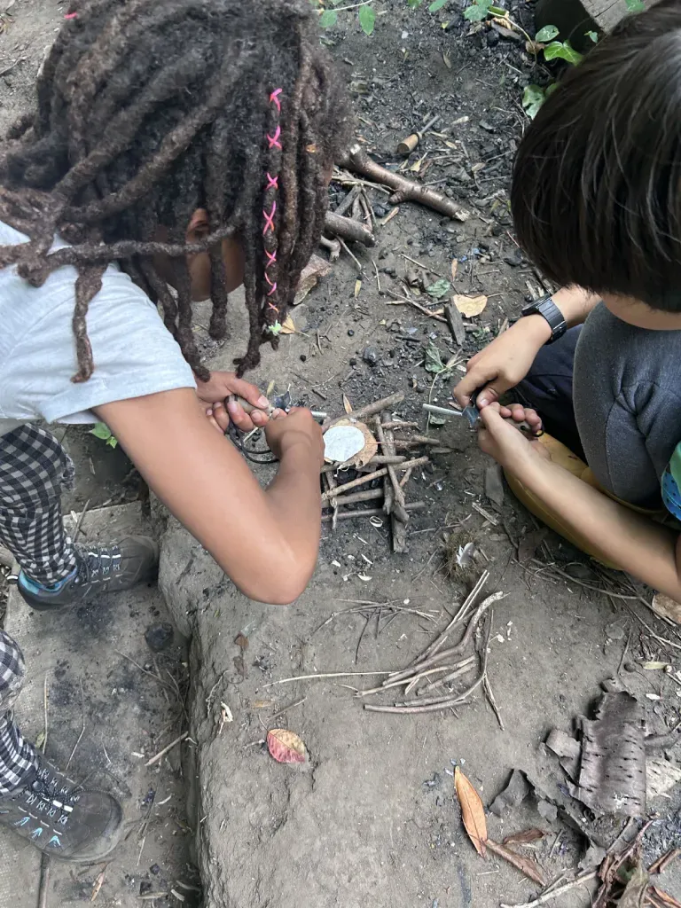Two children crouched together, hands close to a small bow-drill fire kit on the woodland floor