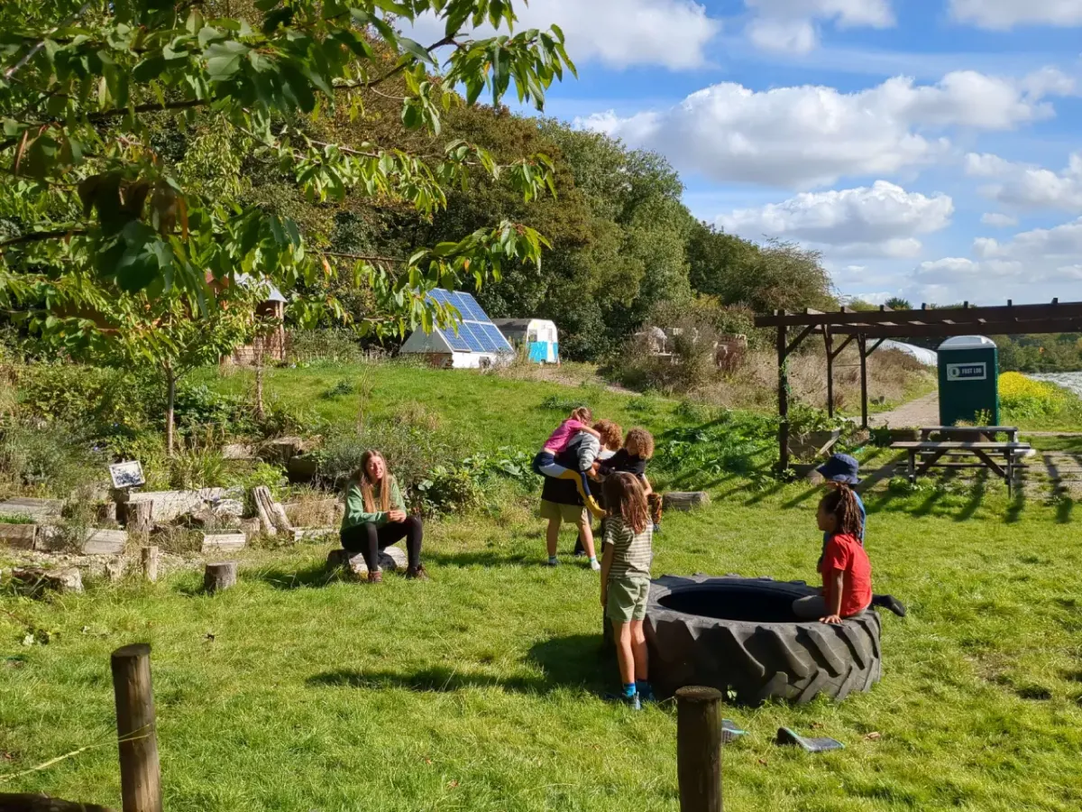 A practitioner sitting on a log while children play together in bright autumn sunshine near a tractor tyre
